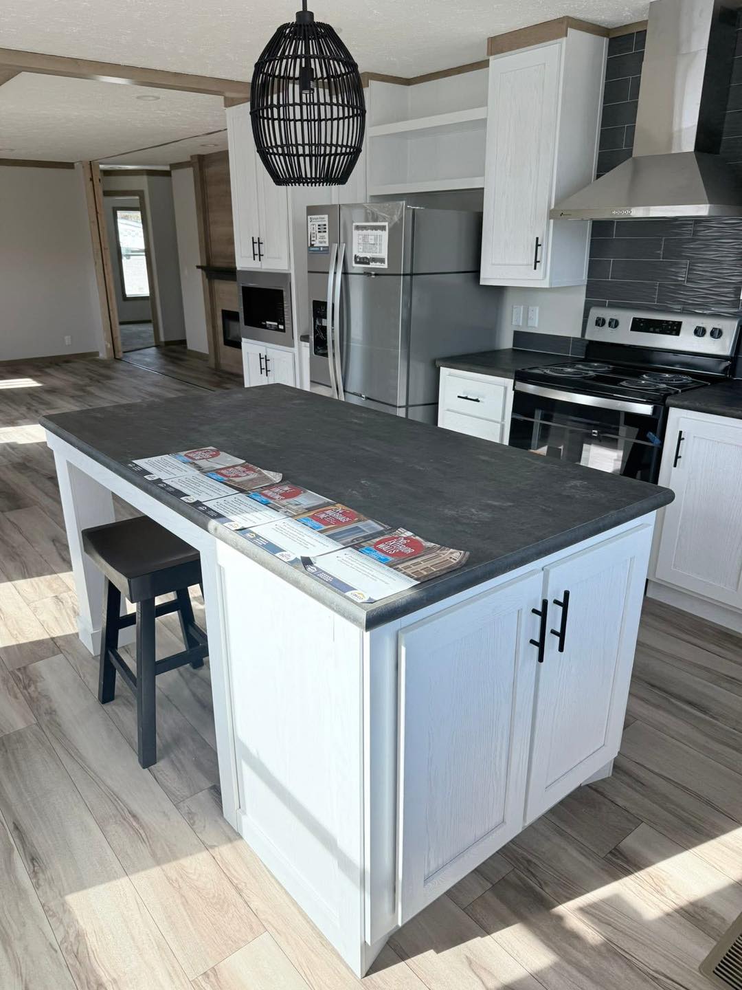 Modern kitchen with white cabinets, a dark stone island, and laminate wood flooring. Stainless steel appliances and a black pendant light enhance the space.