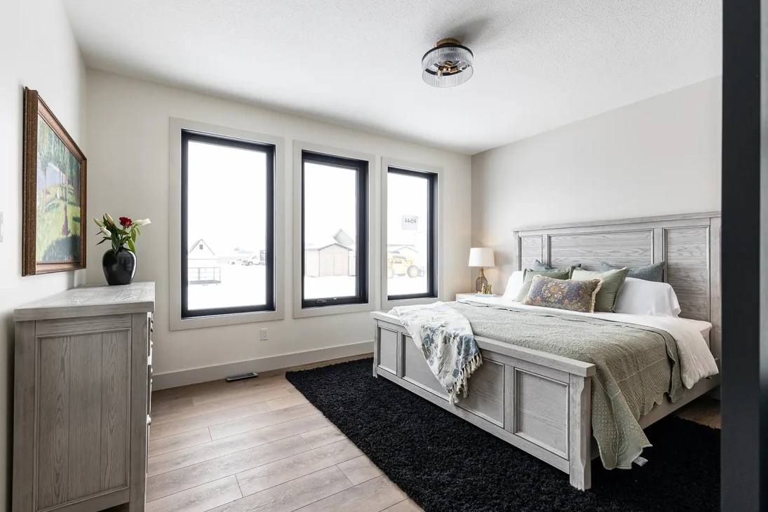 Modern bedroom with a large, gray wooden bed, light bedding, and decorative pillows. Three tall windows, black rug, wooden floor, and a floral artwork on a dresser add elegance.