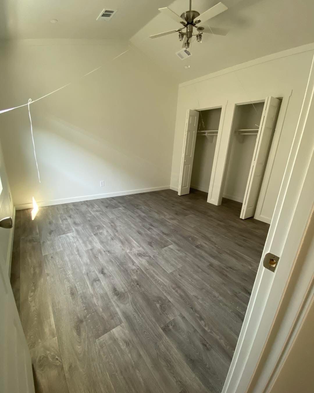 Empty room with gray wood flooring, white walls, two open closets, and a ceiling fan. Sunlight creates a peaceful atmosphere.
