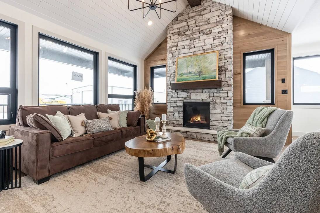 Cozy living room with a high ceiling, featuring a large stone fireplace, brown sofa, gray chairs, rustic coffee table, and large windows.