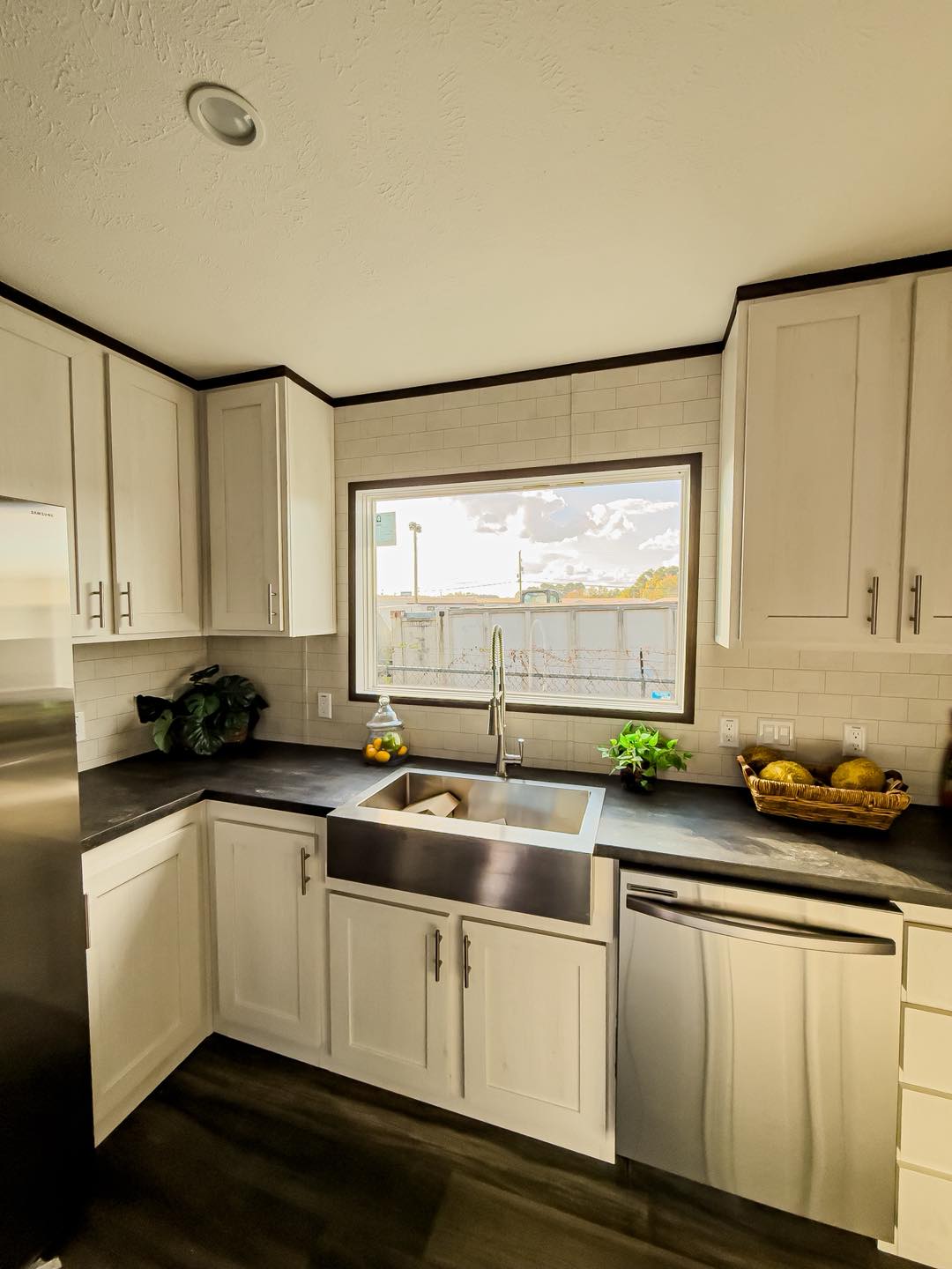 Bright kitchen with white cabinets, a stainless steel farmhouse sink, dark countertops, and a large window. Fresh lemons, greens, and a basket of squash add a vibrant touch.