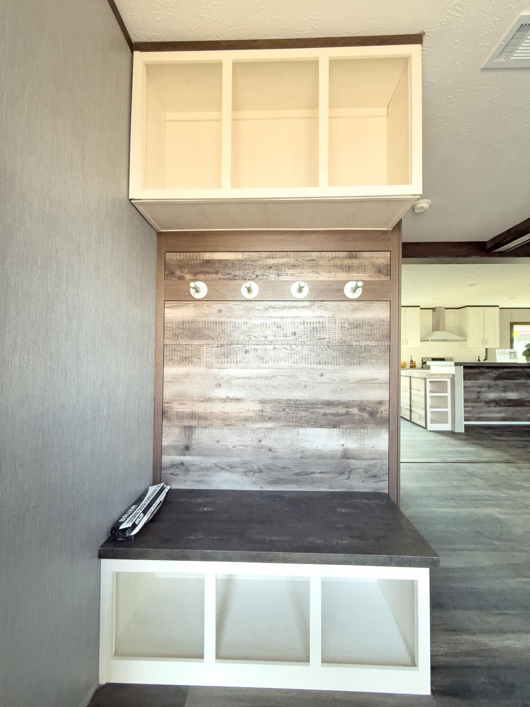Entryway mudroom with gray wood panels, four wall hooks, and storage cubbies above and below a dark bench. Modern kitchen is visible in the background.