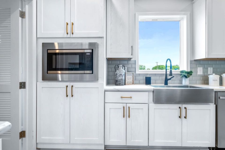 Bright kitchen with white cabinets and brass handles. Features a built-in microwave, large stainless steel sink, and a window with a scenic view.