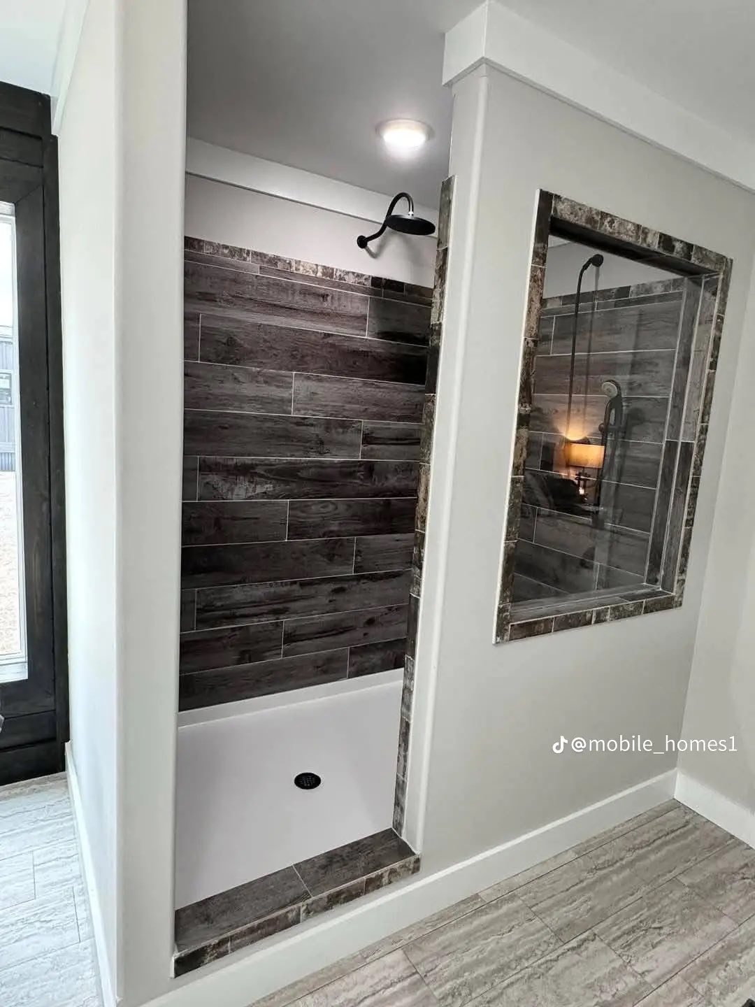 Modern shower with brown wood-like tiled walls and a black rain showerhead. Adjacent window with marble trim, lighted interior creates a cozy feel.