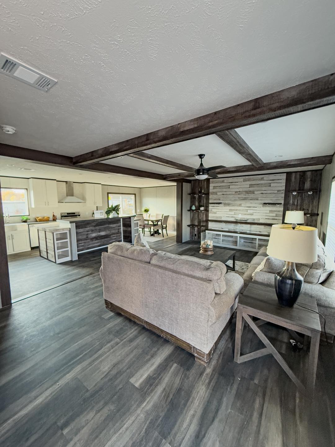Stylish modern living room with gray sofas, dark wood floor, and a contemporary kitchen in the background. It conveys a warm, inviting tone.