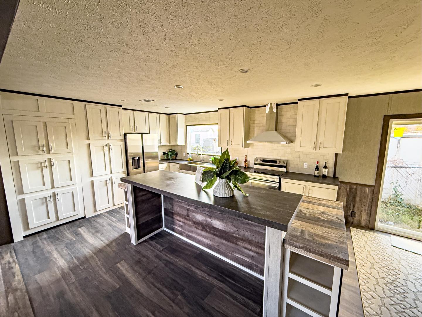 Spacious modern kitchen with dark wood floors, white cabinets, and stainless steel appliances. A large island with a plant centerpiece is central.