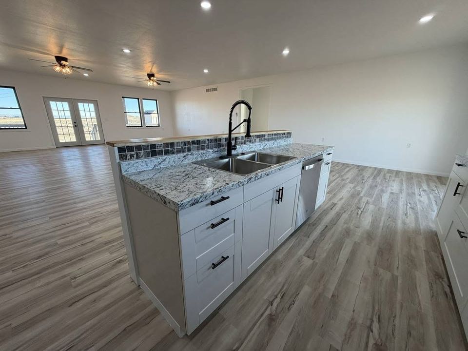 Spacious, modern kitchen with marble countertops, dual sink, and white cabinets in an open-concept room. Hardwood floors and ceiling fans add warmth.