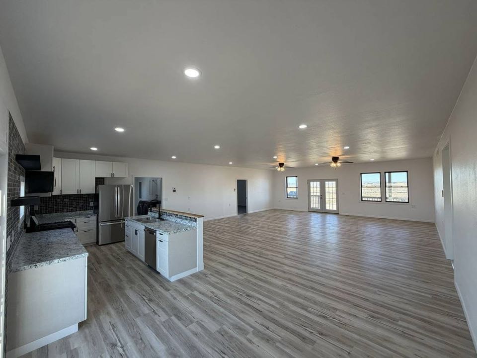 Spacious open-plan kitchen and living area with light wood floors and white walls. Features modern stainless steel appliances and large windows.