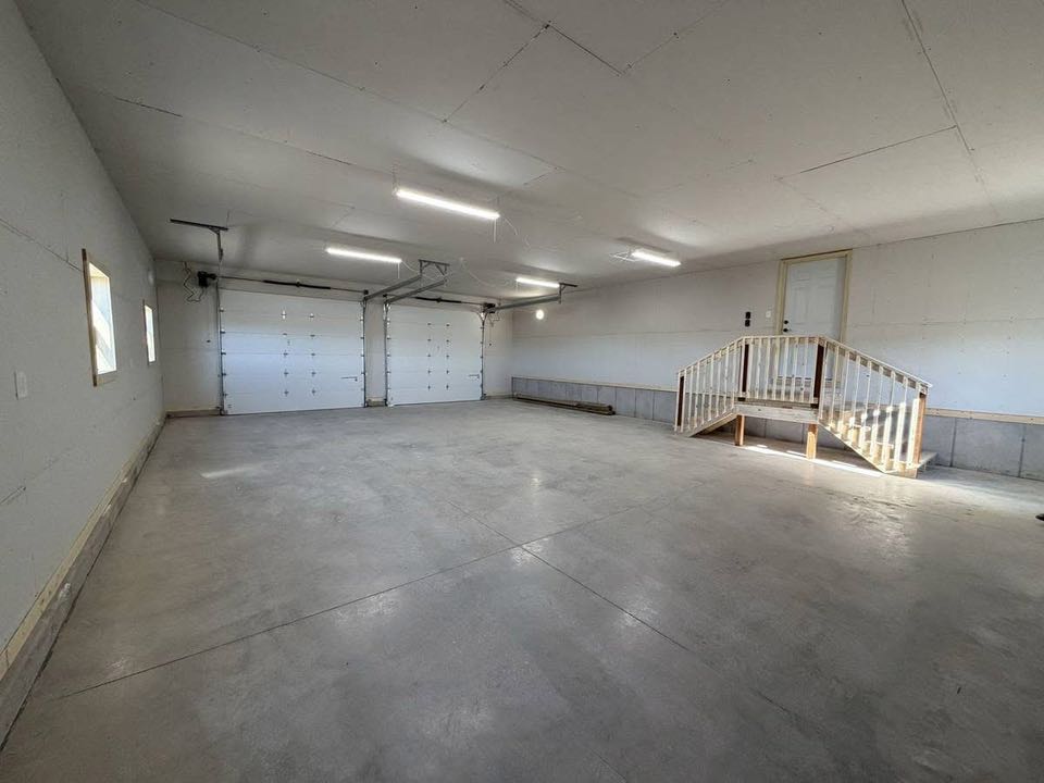 Spacious, empty garage with clean concrete floors, white walls, and bright overhead lighting. Two large garage doors and a wooden staircase lead to a small door.