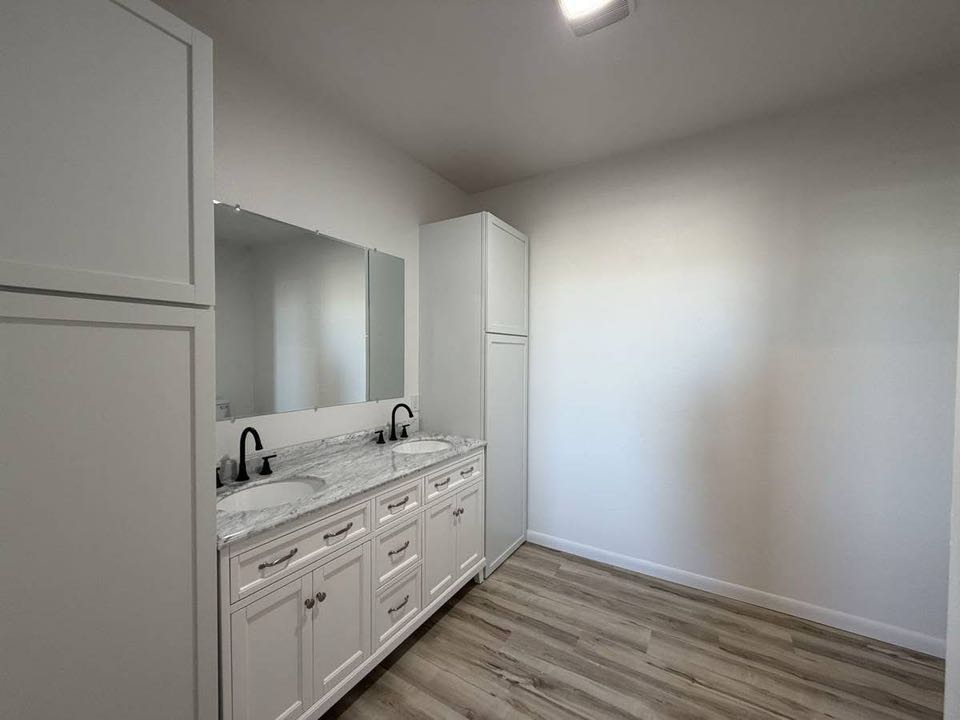 A minimalist bathroom with a white double sink vanity, marble countertop, large mirror, and tall cabinet on light wood flooring, conveying simplicity.