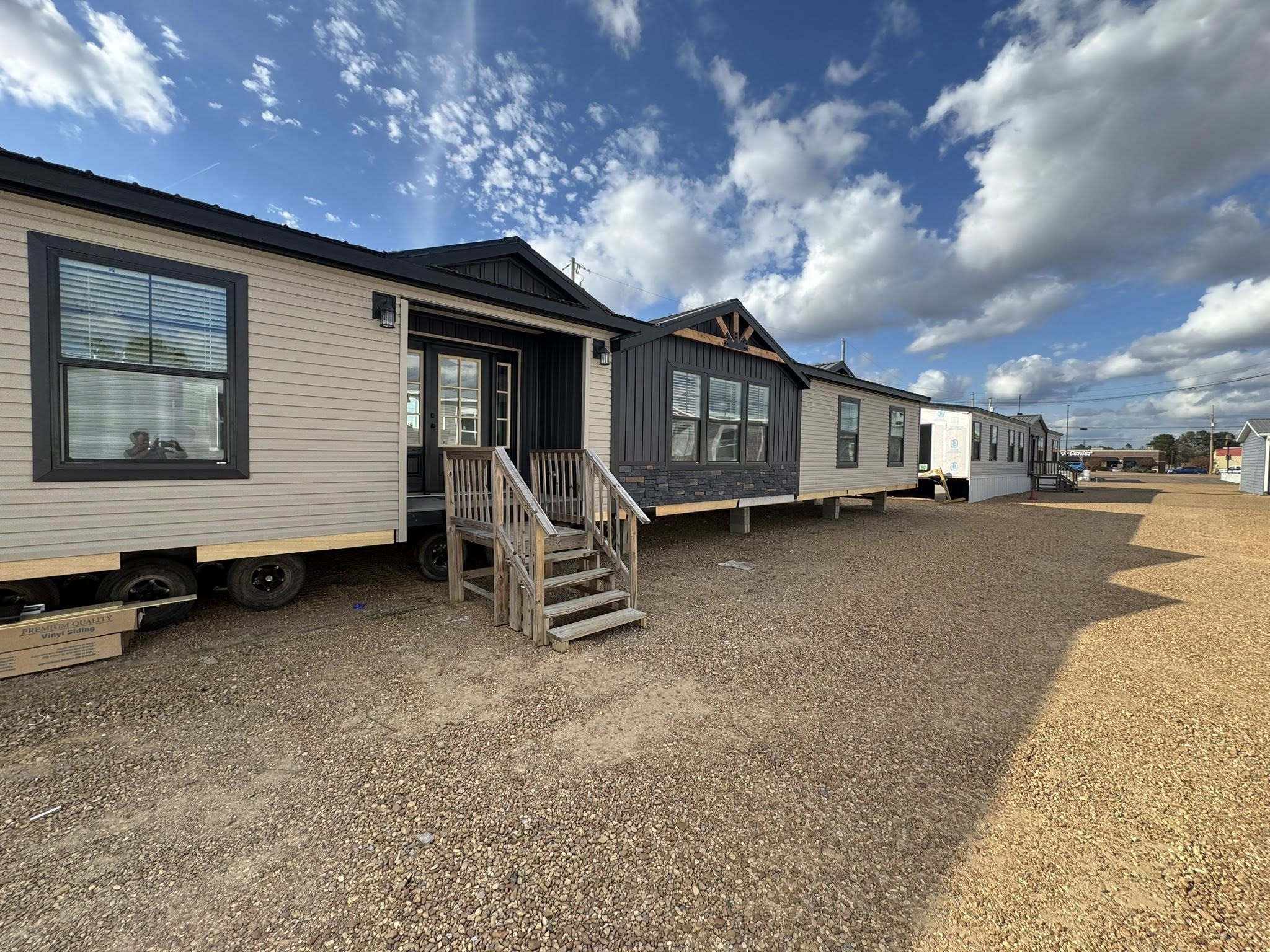 Row of mobile homes on a gravel lot under a partly cloudy sky, conveying a sense of calm and modest living. Simple wooden steps lead up to each unit.