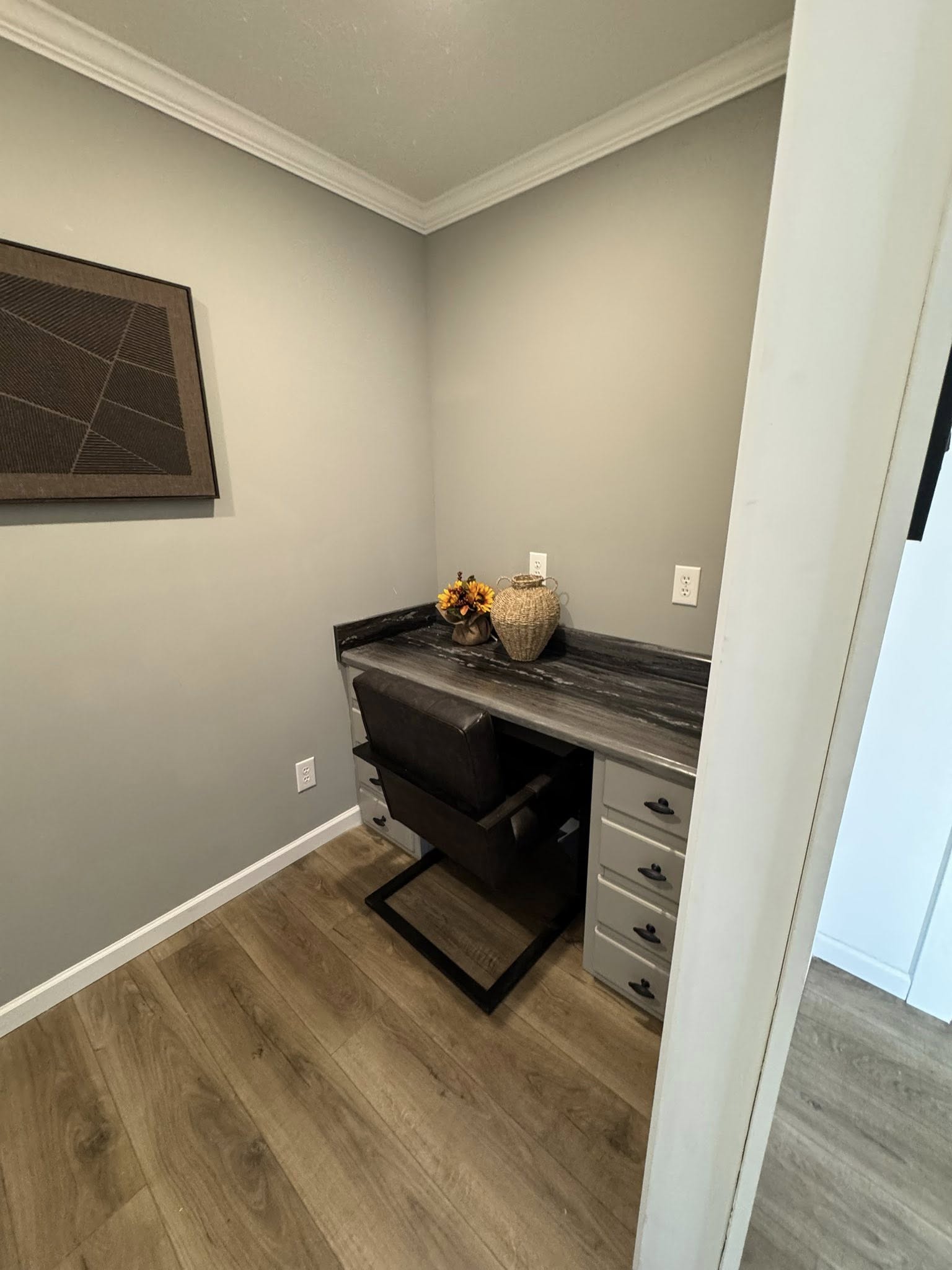 Cozy home office nook with a dark wood desk, gray drawers, and a black chair. Decor includes a basket, sunflowers, and abstract wall art. Warm, inviting tone.