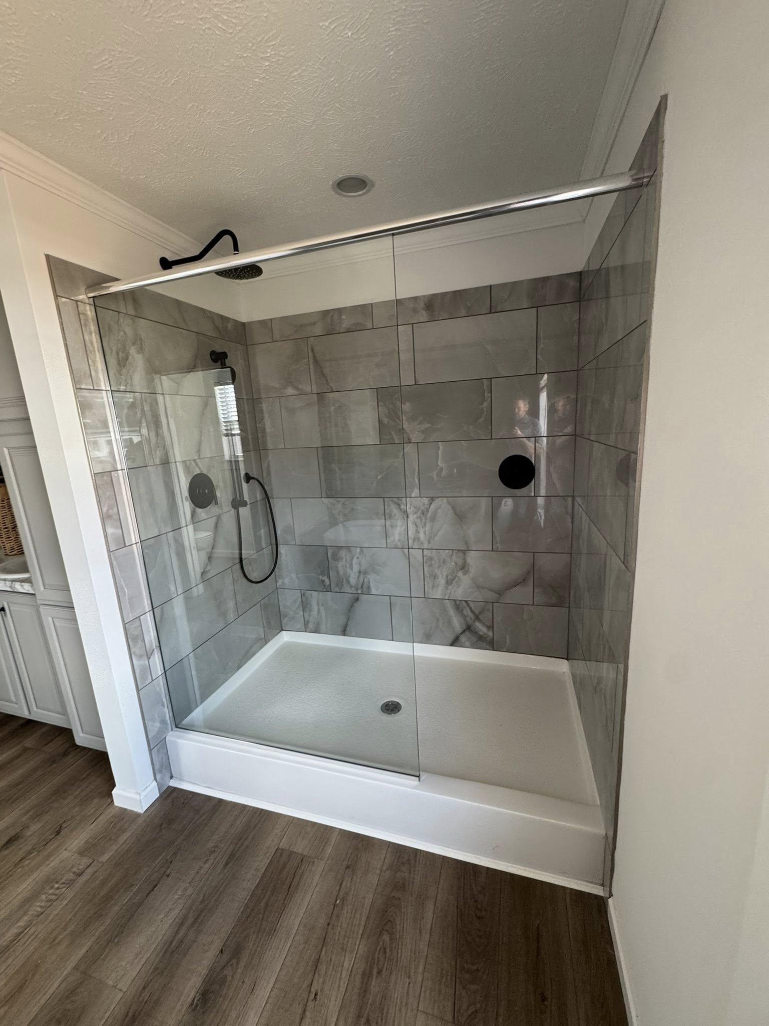 Modern shower with glass doors and gray tile walls. Features a rainfall showerhead and handheld sprayer. Wood-look flooring adds warmth.