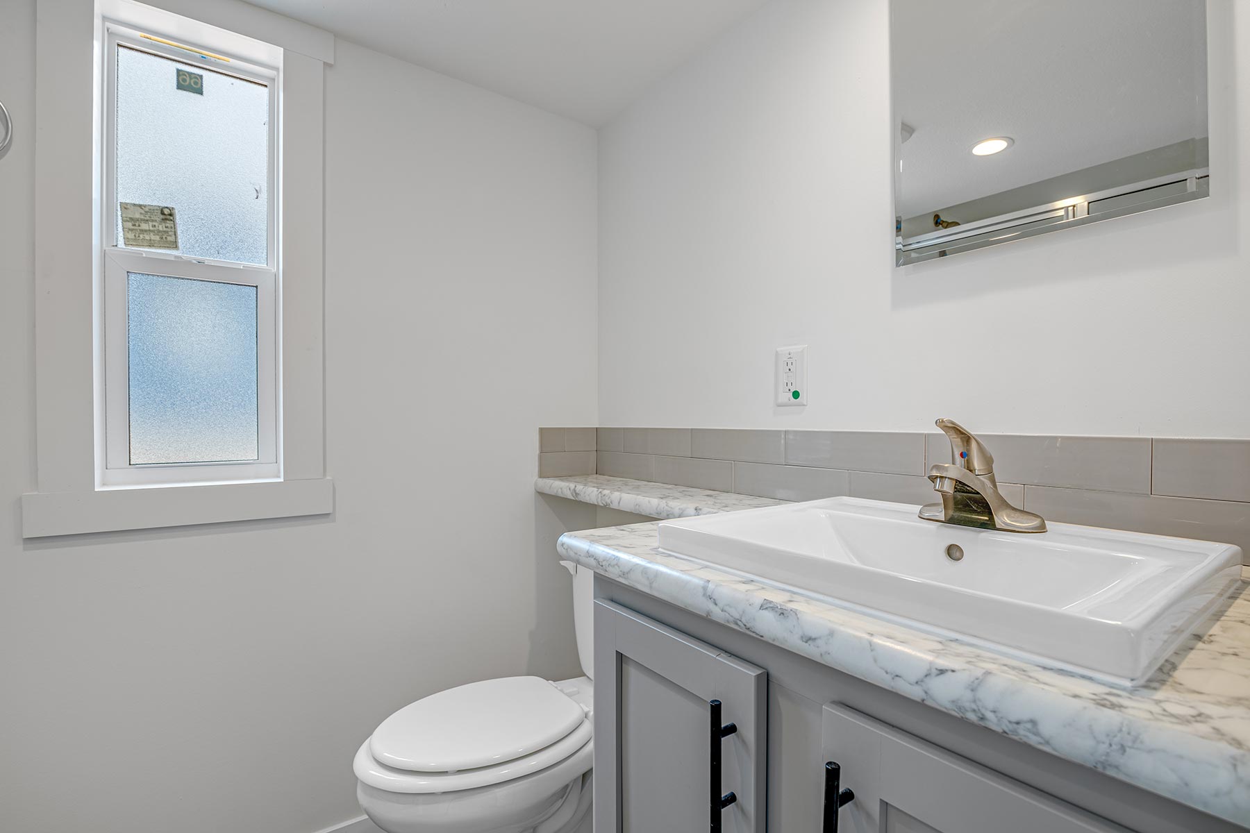 A small, modern bathroom features a white toilet and a marble-topped vanity with a faucet. A frosted window lets in light, enhancing the clean, minimal look.