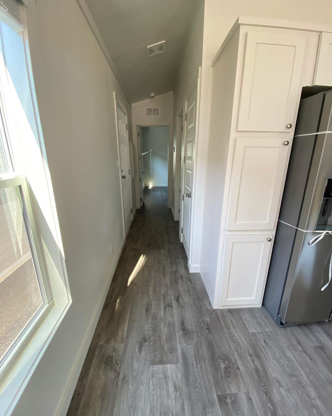 Narrow hallway with light gray walls, wooden flooring, and a bright window to the left. Cabinets and refrigerator are on the right, creating a cozy, modern feel.