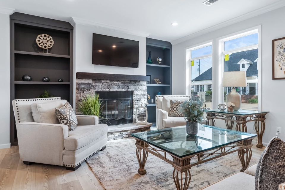 Modern living room with beige armchairs, glass coffee table, and stone fireplace under a mounted TV. Light floods in through large windows.