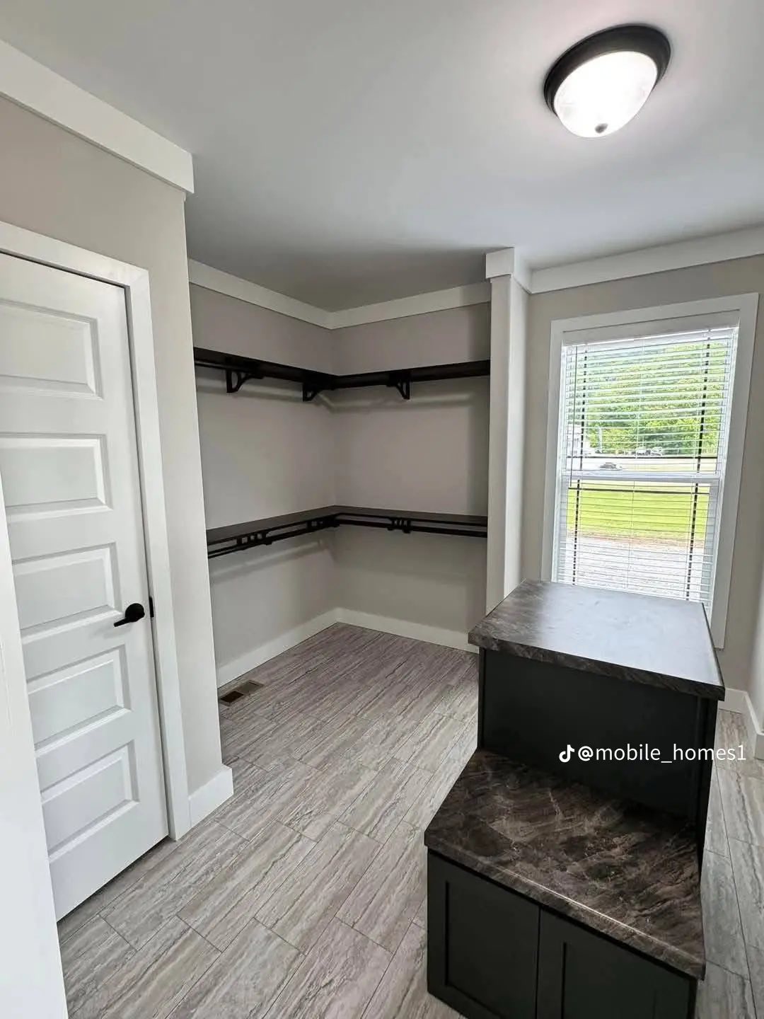Bright, modern walk-in closet with light gray walls, tiled floor, white door, shelving along walls, and a window with blinds offering natural light.