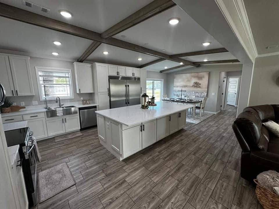 Spacious modern kitchen with white cabinets, island, stainless steel appliances, and wooden beams. Dining area in the background, calm and inviting atmosphere.