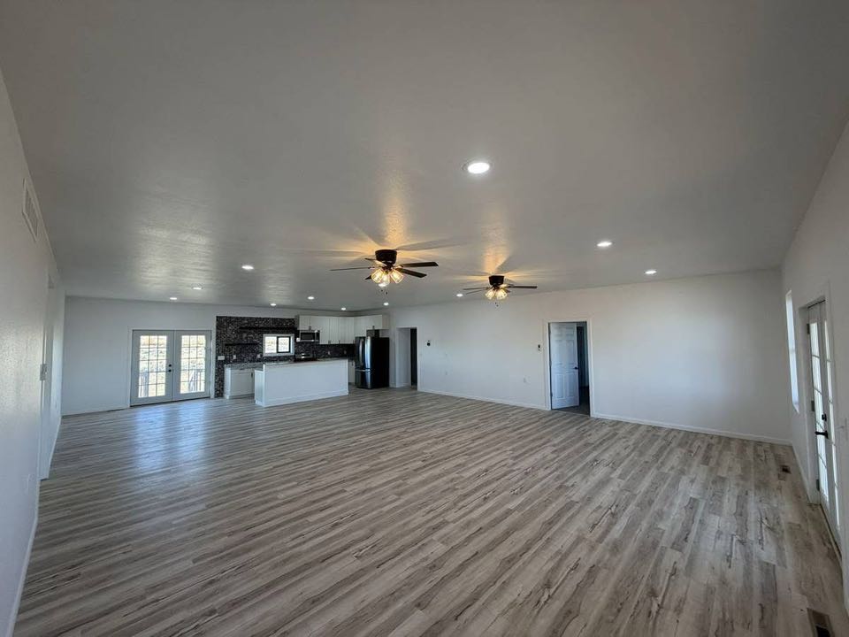 Spacious open-concept room with light wood flooring, white walls, and modern ceiling fans. A kitchen with dark cabinets and stainless appliances. Bright, airy ambiance.