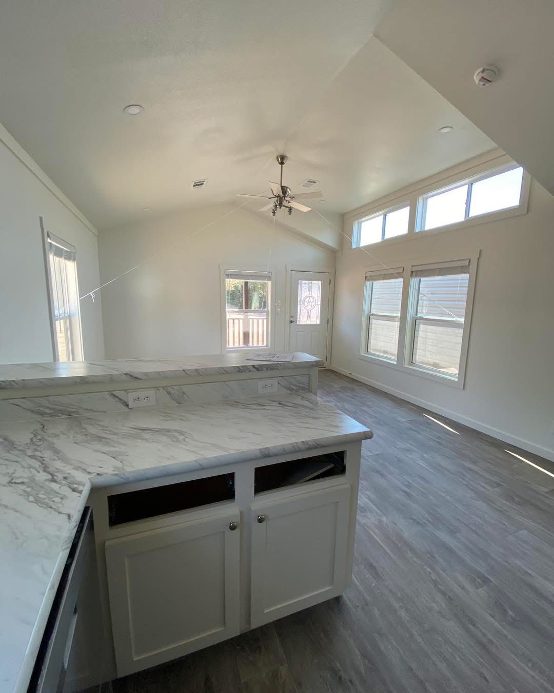 Spacious kitchen with marble countertops, white cabinets, light wood flooring, and large windows. Ceiling fan adds a modern touch. Bright and airy.