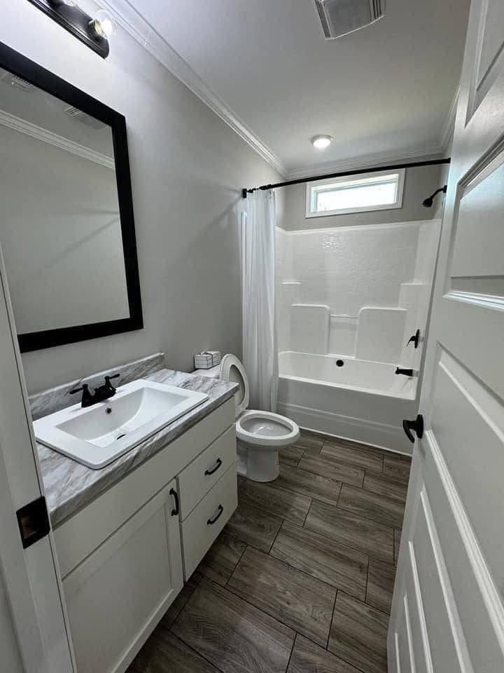 A modern bathroom with a marble-topped vanity, black-framed mirror, and wooden floor. A white bathtub with a curtain is next to a small window.