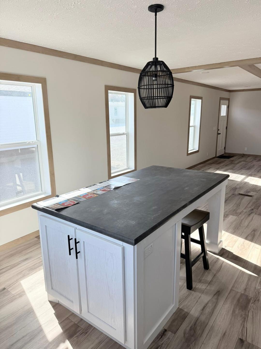 Modern kitchen with a white island, gray countertop, and a single black stool. Sunlit windows, wood floors, and a black woven pendant light create a cozy, bright atmosphere.