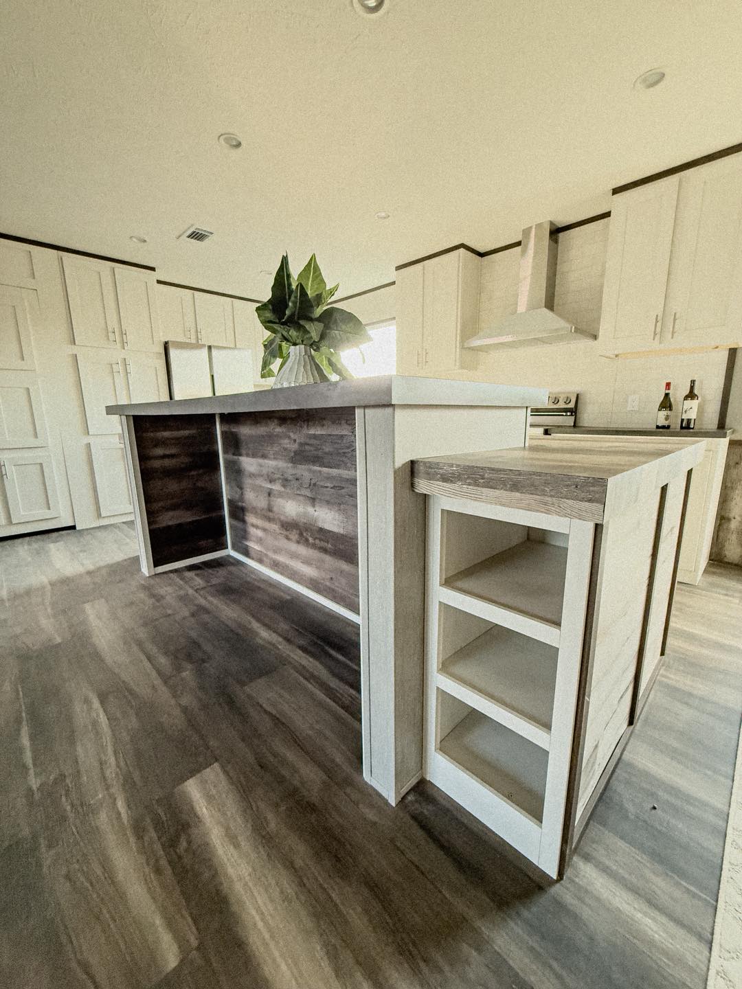 Modern kitchen interior with a large island featuring wood accents and open shelves. White cabinets, a range hood, and a vase with green leaves add elegance.