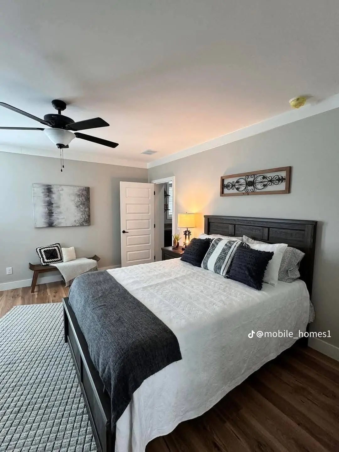 Cozy bedroom with a dark wood bed, white and navy bedding, gray walls, and stylish decor. A ceiling fan and a table lamp add warmth and comfort.