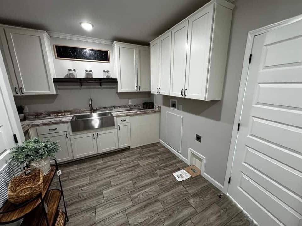 Laundry room with white cabinets, stainless steel sink, and wood-style tile flooring. A shelf with jars and a "Laundry" sign adds charm.