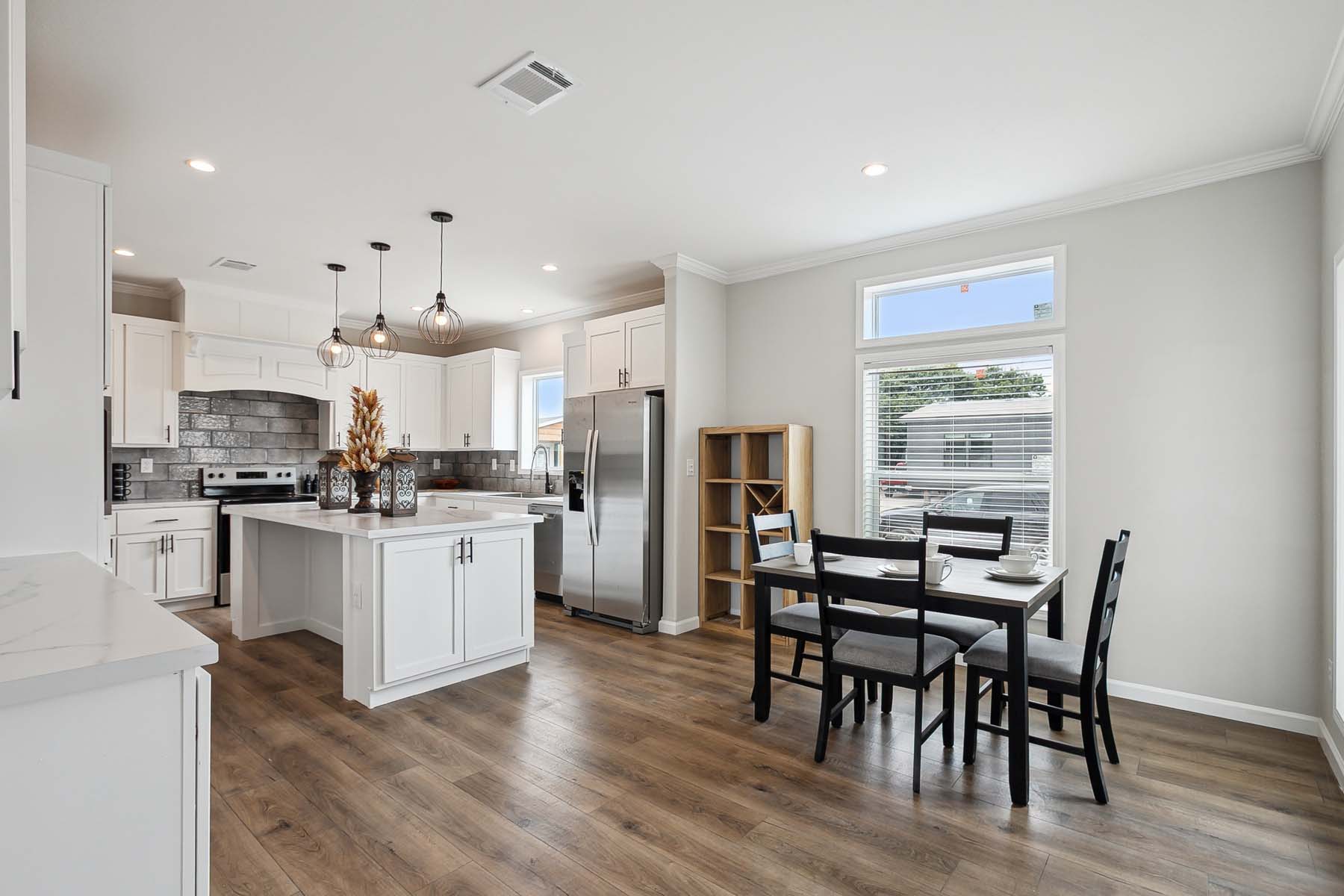 Modern kitchen with white cabinets, marble countertops, and stainless steel appliances. A wooden dining table set for four sits near large windows.