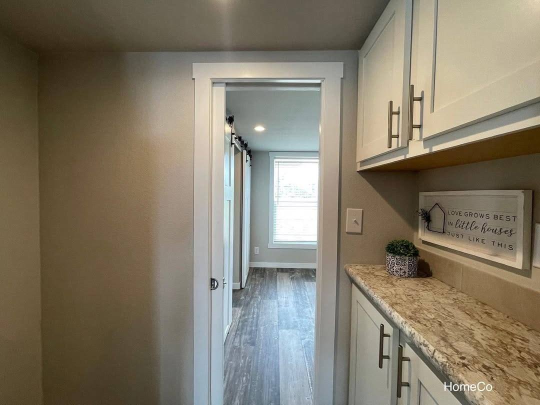 Bright hallway with wood flooring leads to a room with a large window. White cabinet on the right has a decorative sign and plant, creating a cozy feel.