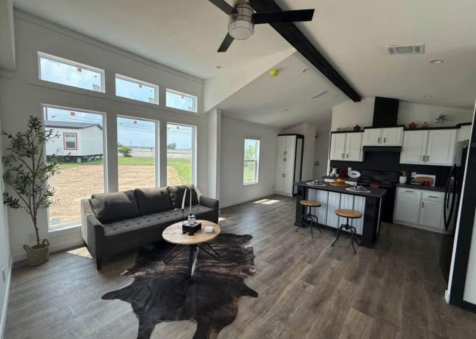 Modern living room and kitchen with large windows, a gray sofa, and a cowhide rug. The kitchen features an island with stools and white cabinets.