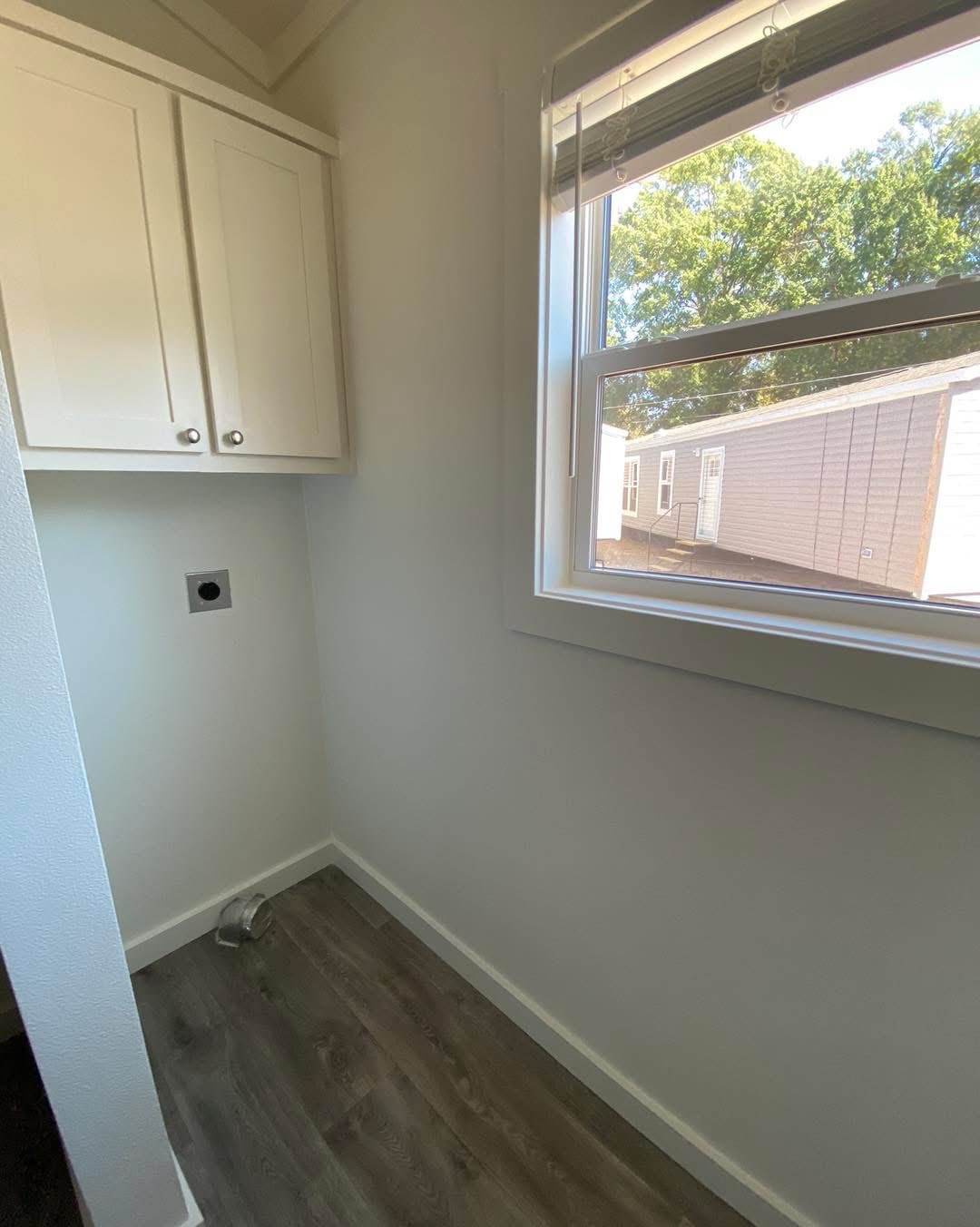 Small laundry room with white cabinets, a window showing a sunny view of trees and neighboring houses, and a flooring outlet for appliances.