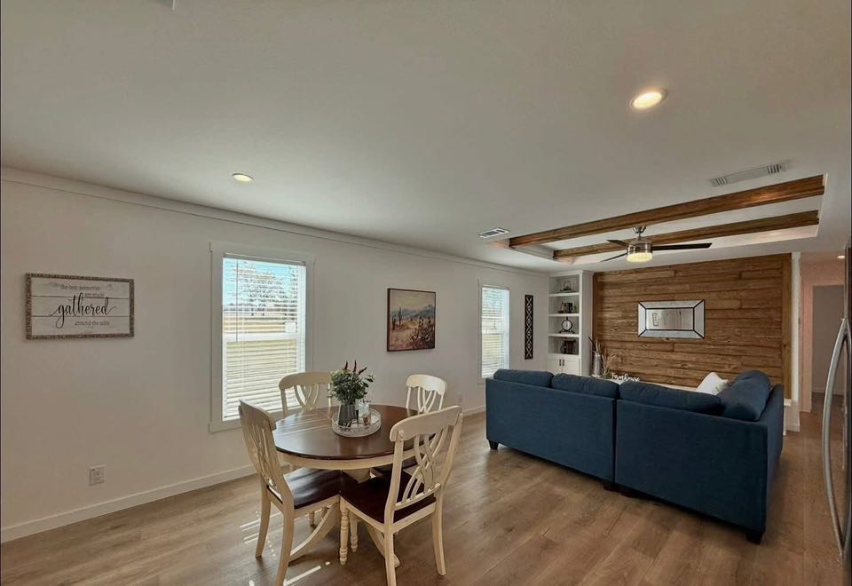 A cozy living room with a round wooden dining table and four chairs on the left. A navy L-shaped sofa faces a wooden accent wall with shelves on the right. Light streams through two windows, creating a warm, inviting atmosphere.