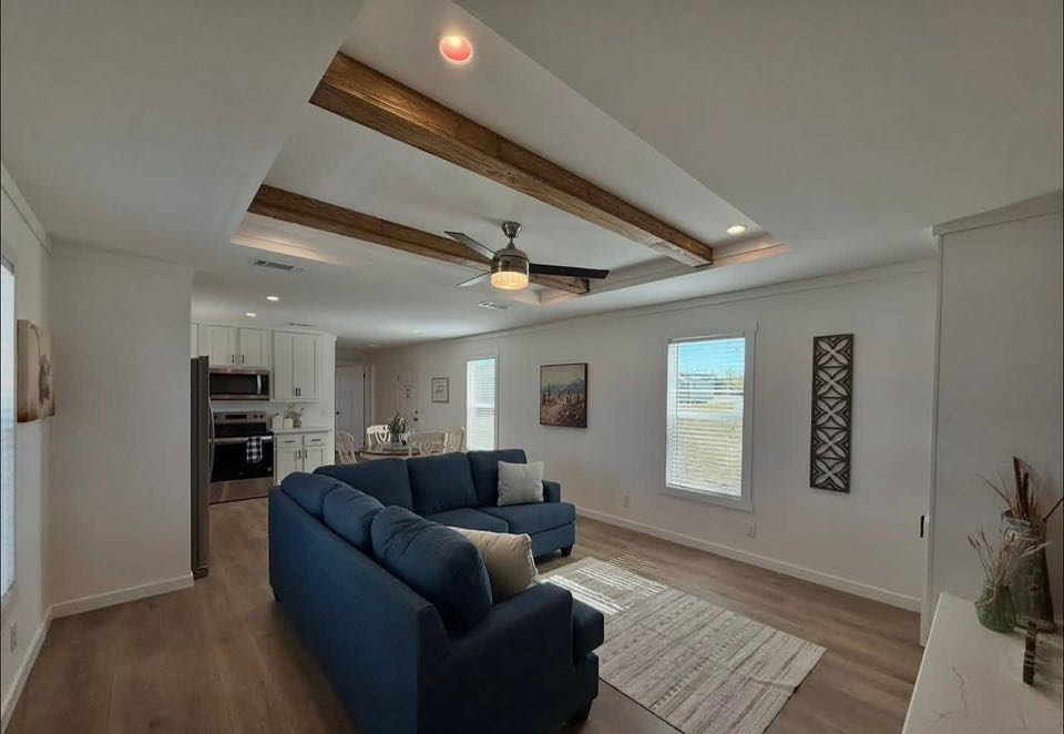 Modern living room with wooden beams features a blue sectional sofa and gray chairs. Light streams through windows, creating a cozy and inviting atmosphere.