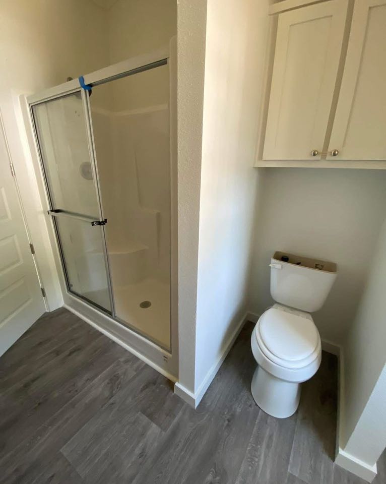 A compact bathroom with a glass shower enclosure on the left and a white toilet on the right. Gray wooden floor adds a modern, clean look.