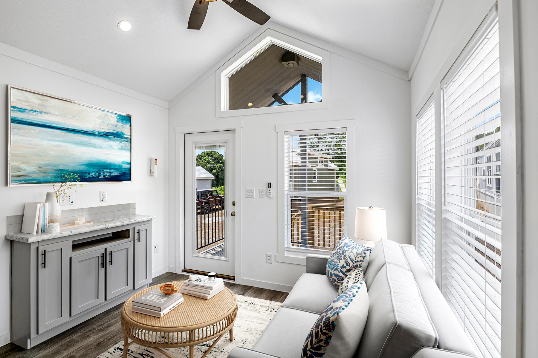 A bright, modern living room with white walls and a vaulted ceiling. Gray couch with patterned cushions, round wicker coffee table, landscape artwork, door, windows with blinds, and a skylight evoke a cozy ambiance.