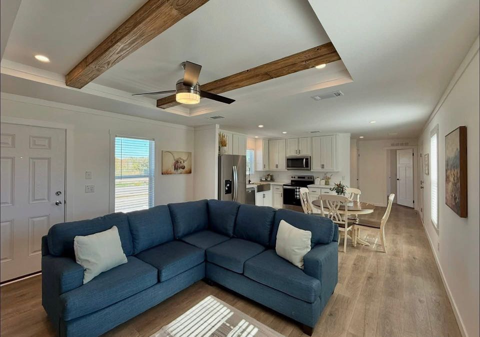 Modern open-concept living area with a blue sectional sofa, wooden ceiling beams, and a kitchen featuring white cabinets and stainless steel appliances. Cozy and inviting.