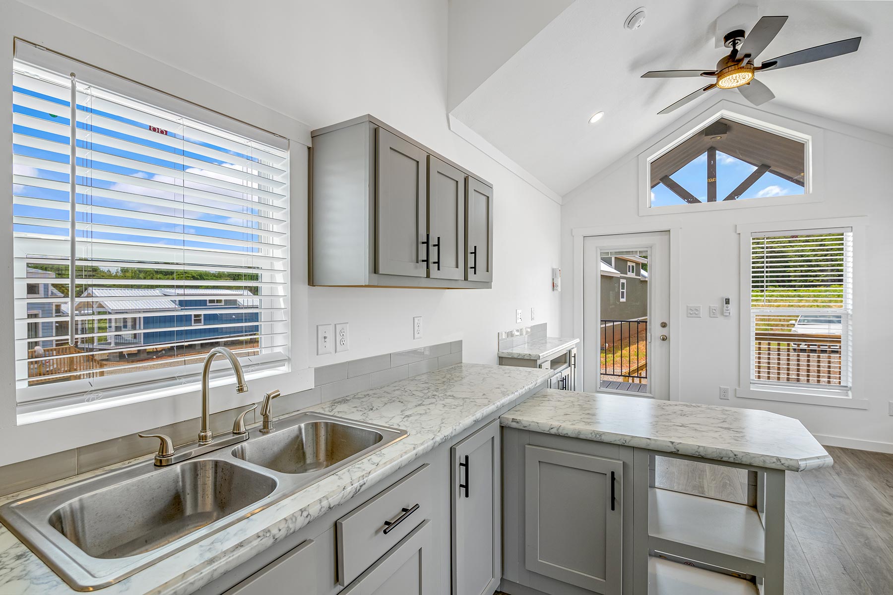 Bright, modern kitchen with gray cabinets, marble countertop, and double sink. Large windows, ceiling fan, and airy atmosphere with natural light.