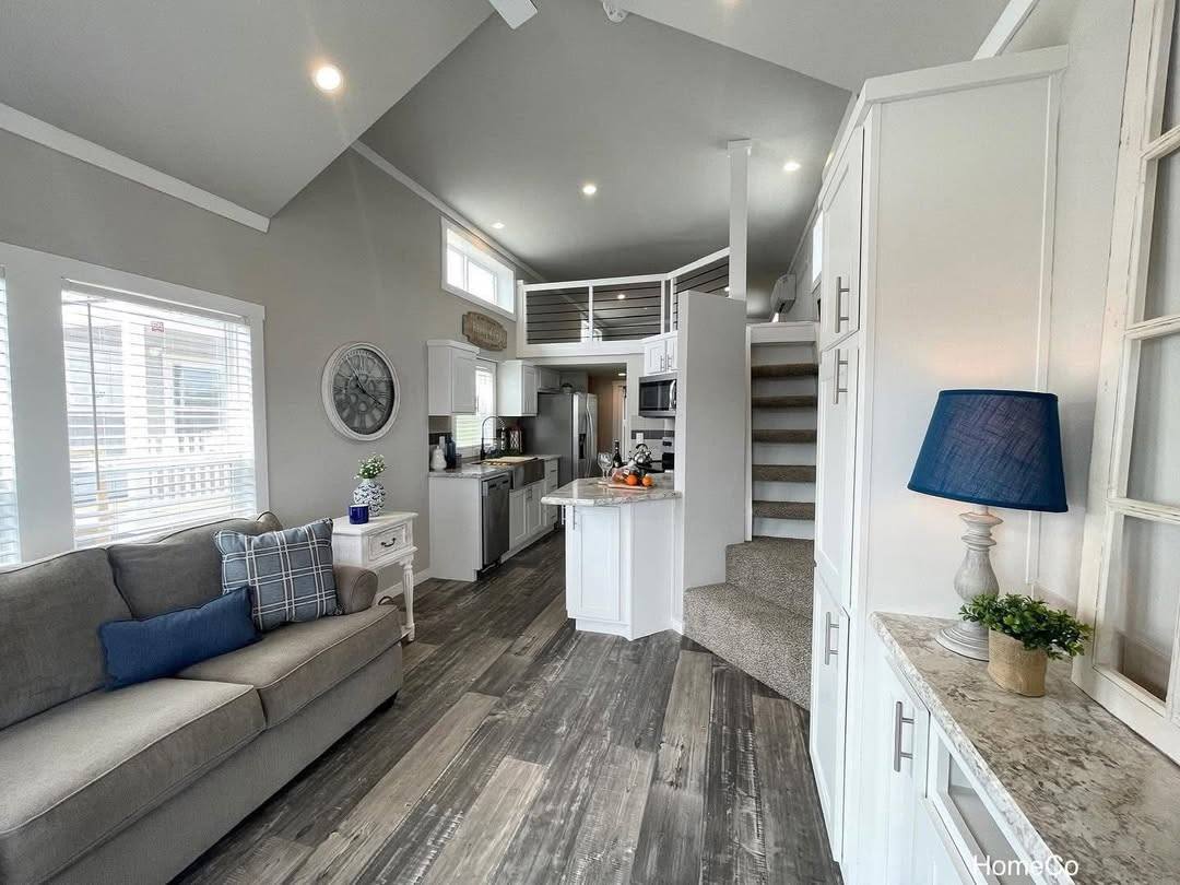 Spacious modern living area with vaulted ceiling, gray sofa, and blue accents. White cabinets, wood flooring, kitchen island, and steps leading to open loft. Cozy ambiance.