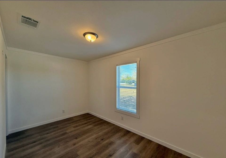 Empty room with white walls and dark wood flooring. A single window with blinds lets in natural light. A ceiling light fixture adds warmth. Minimalistic and serene ambiance.