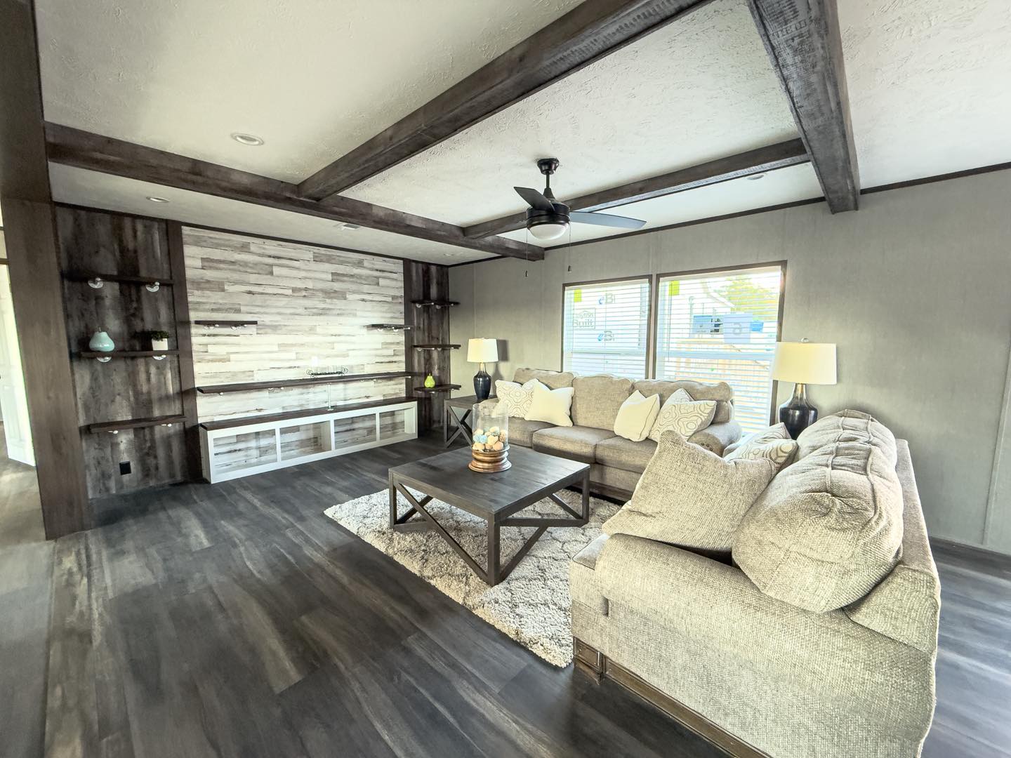Modern living room with gray L-shaped sofa, light wood paneled wall with shelves, dark wood beams, and a ceiling fan. Bright and inviting atmosphere.