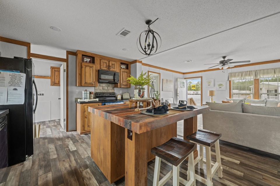Modern kitchen and living room with a rustic wooden island, stools, and dark appliances. Bright room with large windows and ceiling fan. Cozy and inviting.
