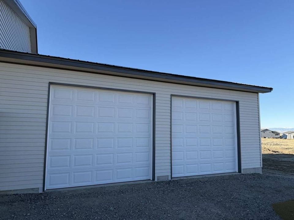 A cream-colored garage with two large white doors is shown under a clear blue sky. The setting is open, with a few distant houses visible.