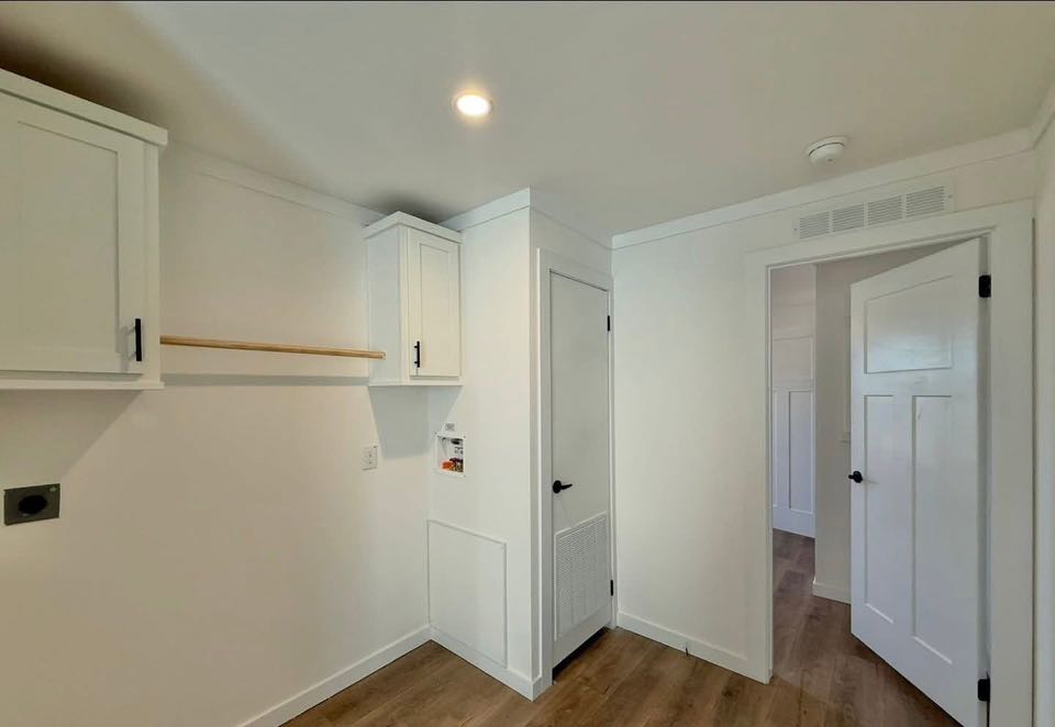Bright utility room with white walls, wooden floor, cabinets, and open door leading to hallway. Mood is clean and minimalist.
