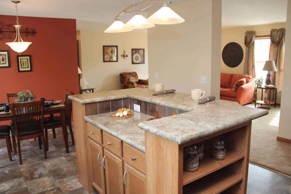 A cozy kitchen with a stone countertop island, wooden cabinets, and pendant lights. Dining area on left with red accent wall; living room with orange couch on right.