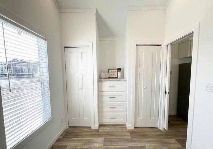 Bright room with white walls and wooden floor, featuring two closets and a central chest of drawers. Decor includes a plant and a “Bless this Nest” sign, evoking a calm, cozy atmosphere.