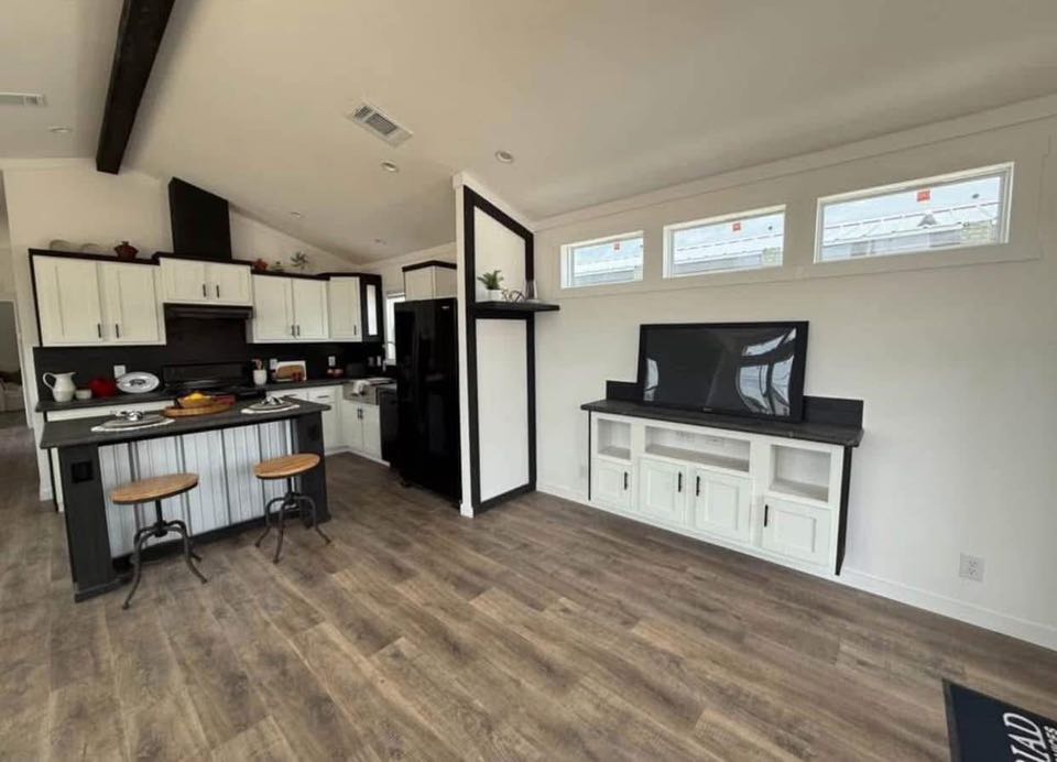 Modern kitchen-living area with white cabinets, black countertops, bar stools, and wall-mounted TV. Bright space with natural wood flooring. Cozy and sleek.