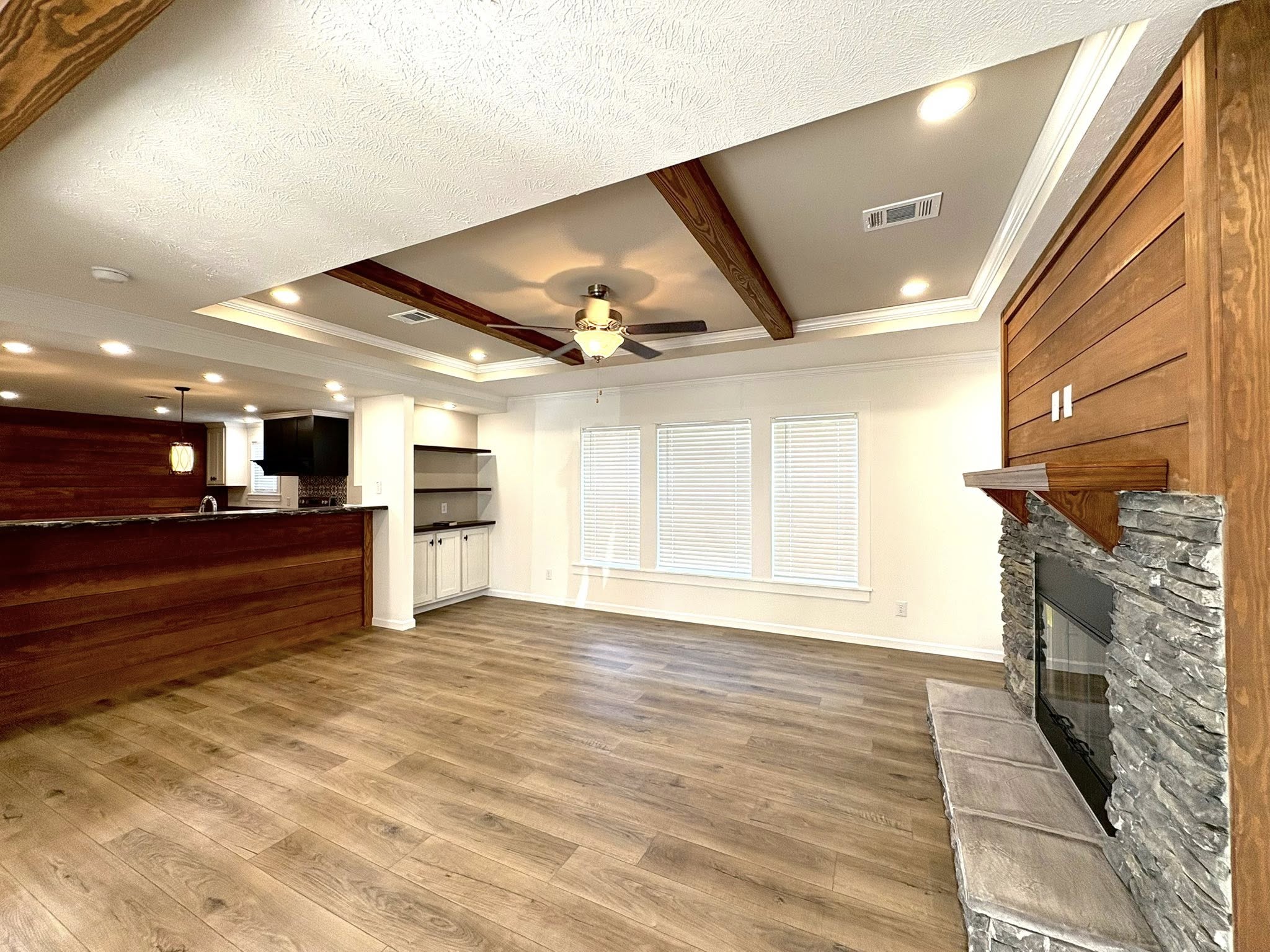 Spacious modern living room with wood beams, stone fireplace, and kitchen bar counter. Warm hardwood floors, recessed lighting, and a ceiling fan offer a cozy, inviting tone.