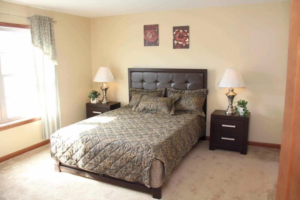 A cozy bedroom with a large bed, brown quilted bedding, and a padded headboard. Two nightstands with lamps and plants flank the bed. Sunlight streams through a window, and two floral paintings decorate the wall, creating a warm, inviting atmosphere.